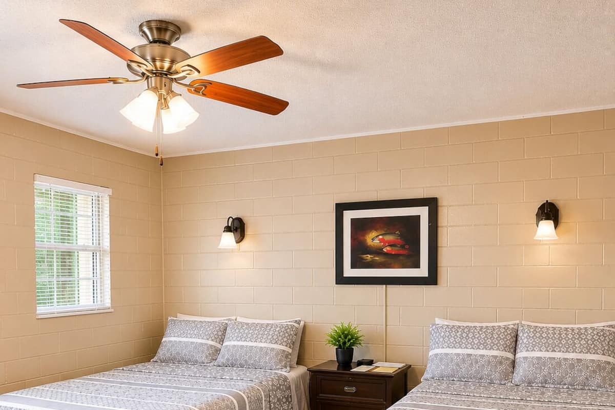A bedroom with two beds, a ceiling fan, a window with blinds, a nightstand, a framed picture on the wall, and a red chair.