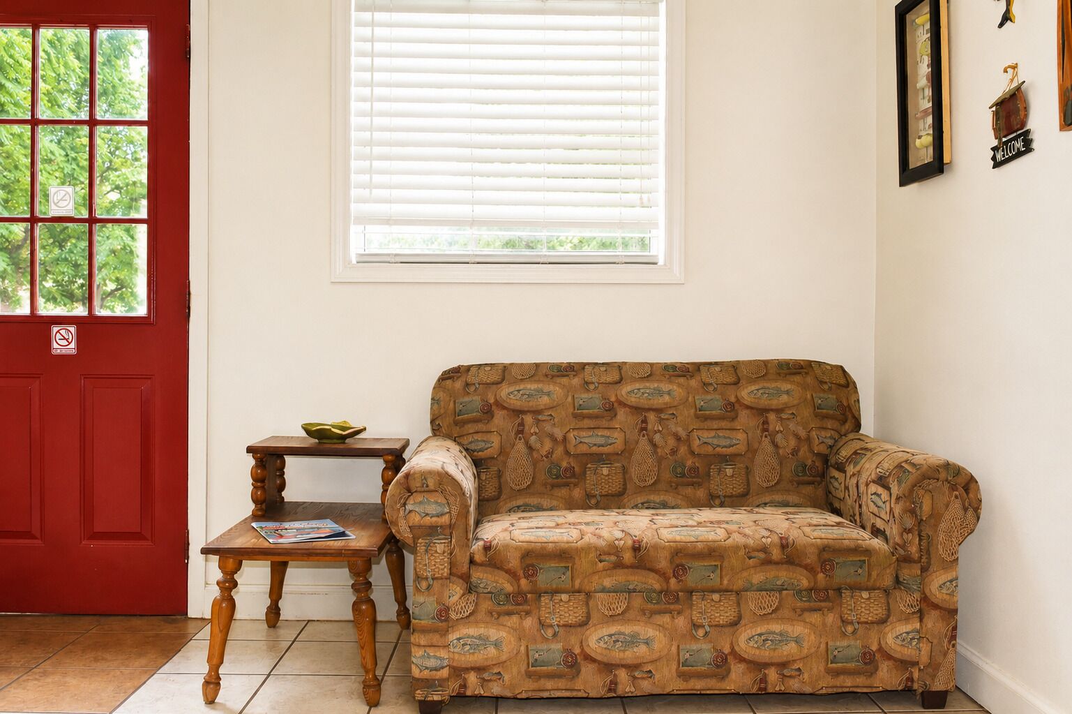 A room with a patterned sofa, a small wooden table, a red door, and a window with blinds.