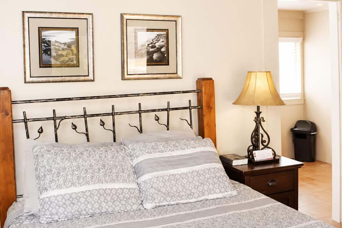 A bedroom with a bed, two framed pictures on the wall, a nightstand with a lamp, and a ceiling fan.