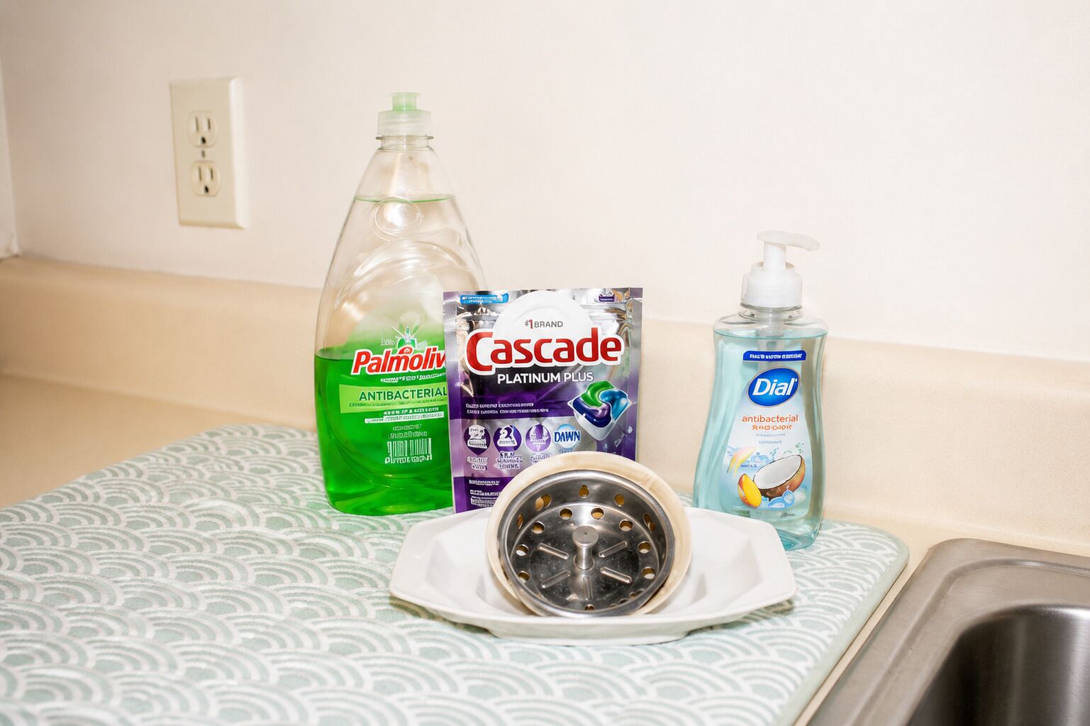 A bathroom sink with a drain cover, a green bottle of Palmolive, a box of Cascade Platinum Plus, and a bottle of Dial antibacterial hand soap.