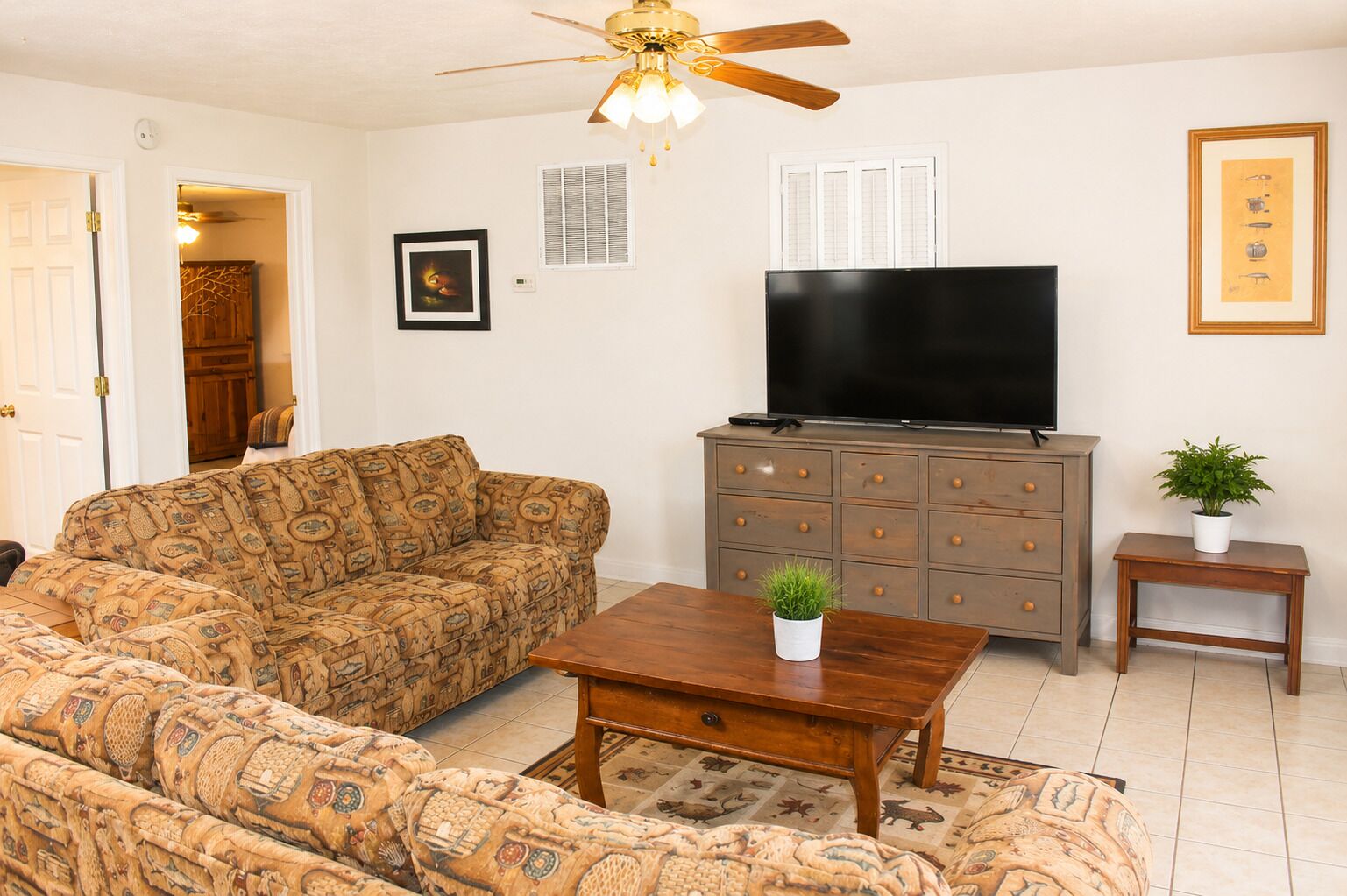 A living room with a sofa, a coffee table, a TV on a wooden cabinet, a ceiling fan, and a framed picture on the wall.