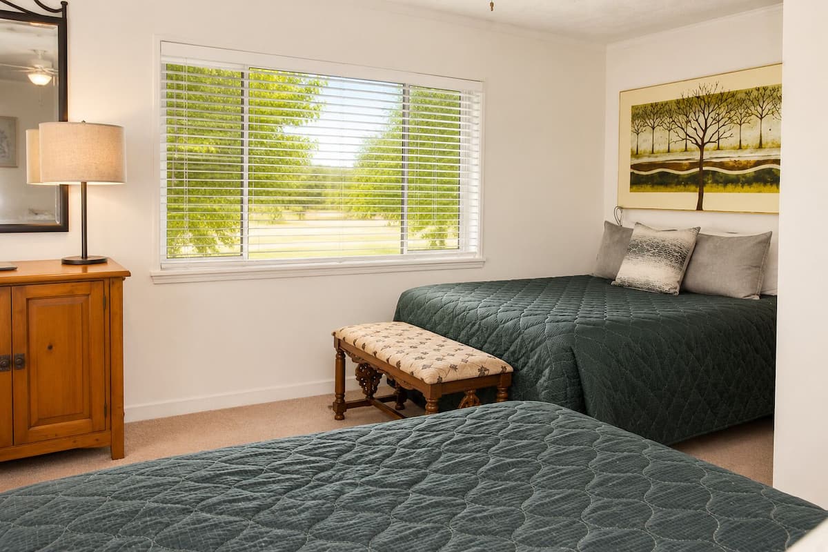 A bedroom with two beds, a bench, a lamp, a window with blinds, and a painting on the wall.