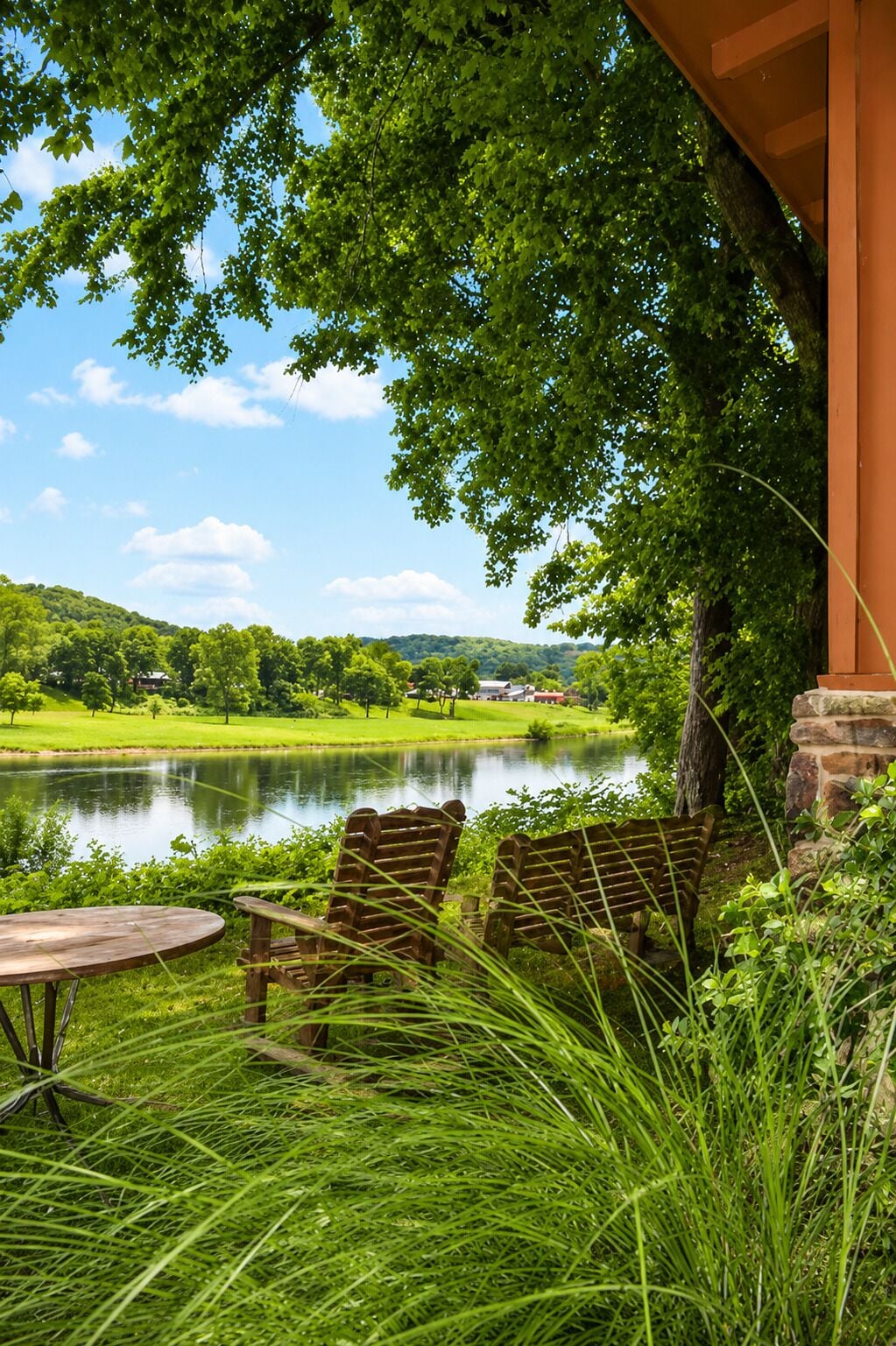 A lakeside patio with wooden chairs and a table, surrounded by lush greenery and a view of a lake and distant hills.