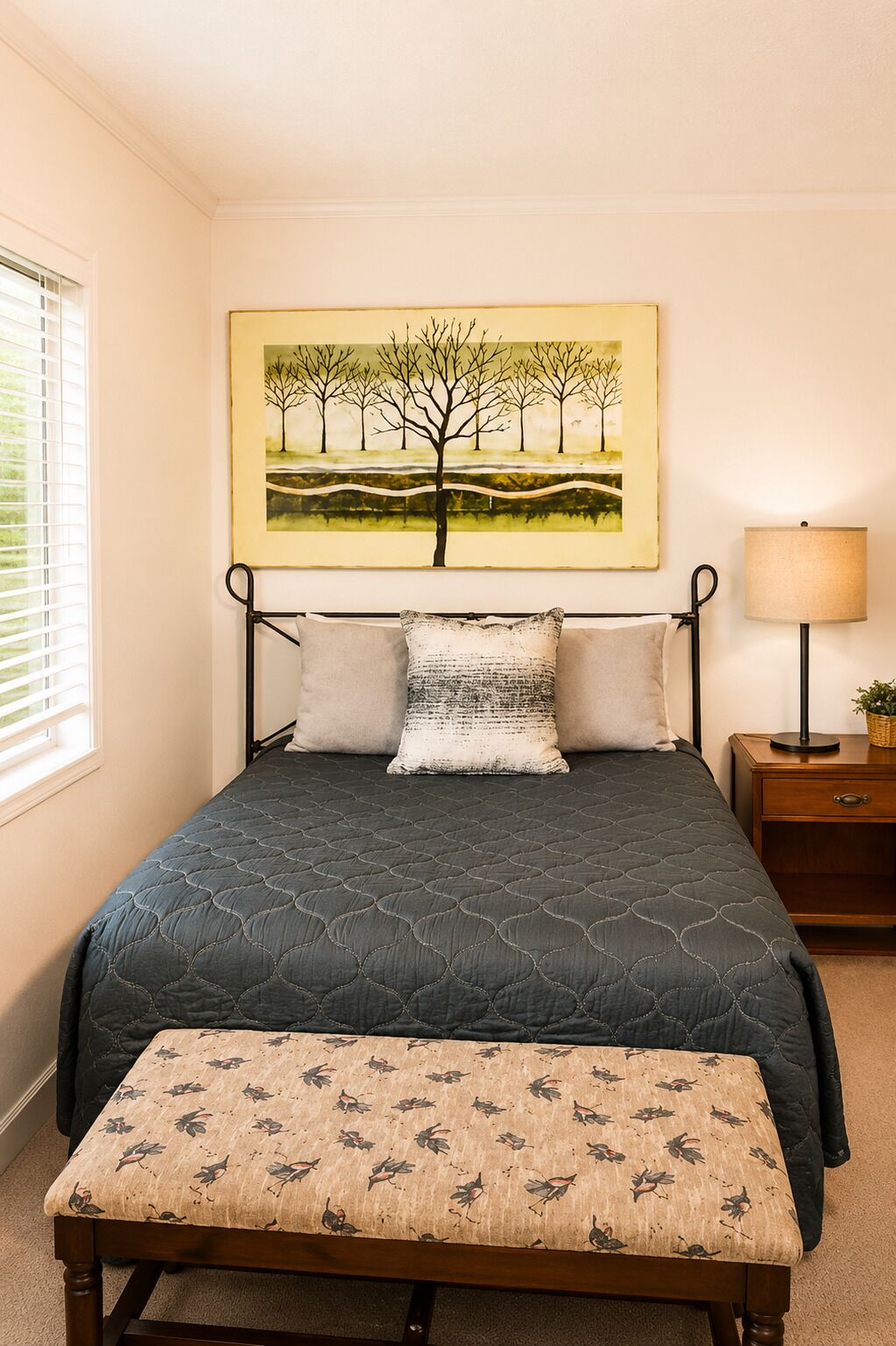 A bedroom with a bed, pillows, a bedside table with a lamp, a bench, and a painting on the wall.
