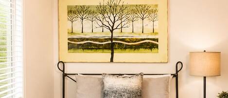 A bedroom with a bed, pillows, a bedside table with a lamp, a bench, and a painting on the wall.