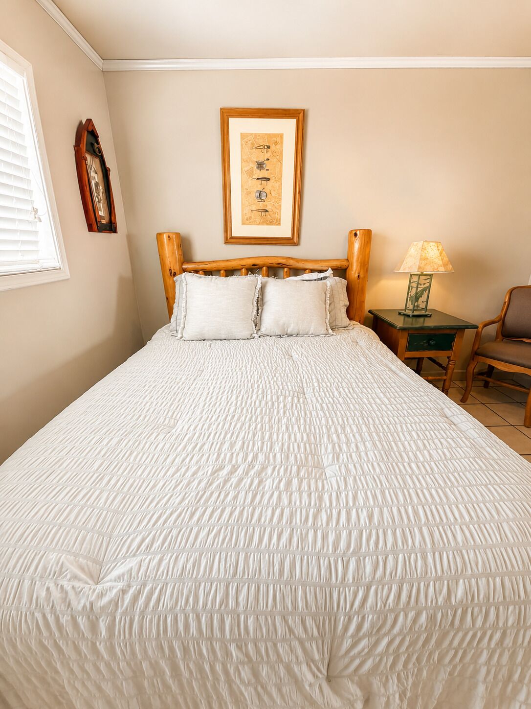 A neatly made bed with a quilted white cover, a wooden headboard, a framed picture on the wall, a bedside lamp, and a chair.