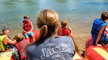 A group of people on a raft with life jackets, a woman in a 'JACKS' shirt, and a river with trees in the background.