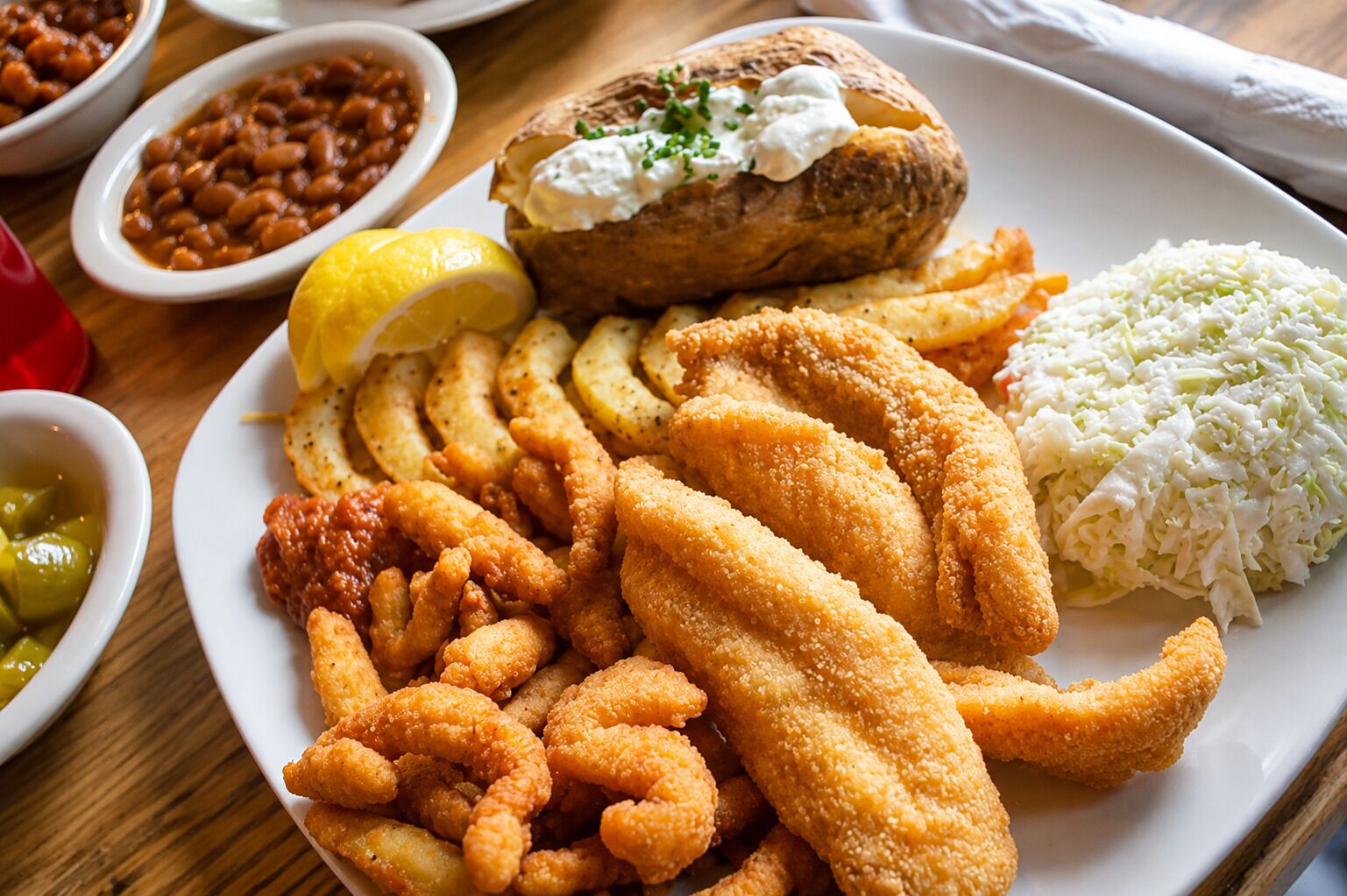 Une assiette de nourriture comprenant du poisson-frites, une pomme de terre au four, de la salade de chou et un quartier de citron.