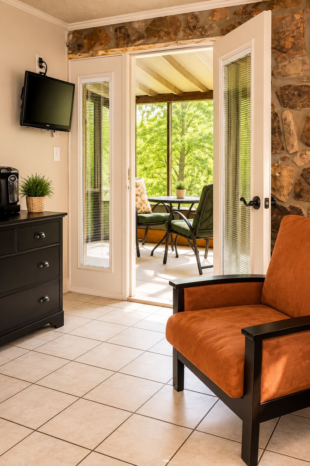 A room with a TV, a coffee maker, a plant, a sofa, and a view of an outdoor patio with a table and chairs.