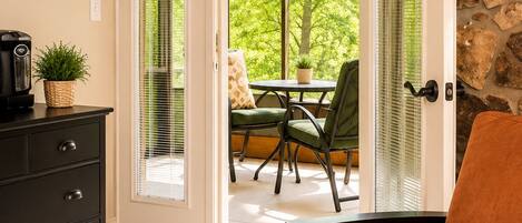 A room with a TV, a coffee maker, a plant, a sofa, and a view of an outdoor patio with a table and chairs.