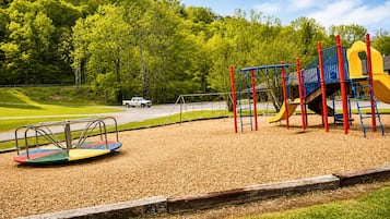 A playground with a swing set, a slide, and a seesaw on a grassy area with trees in the background.