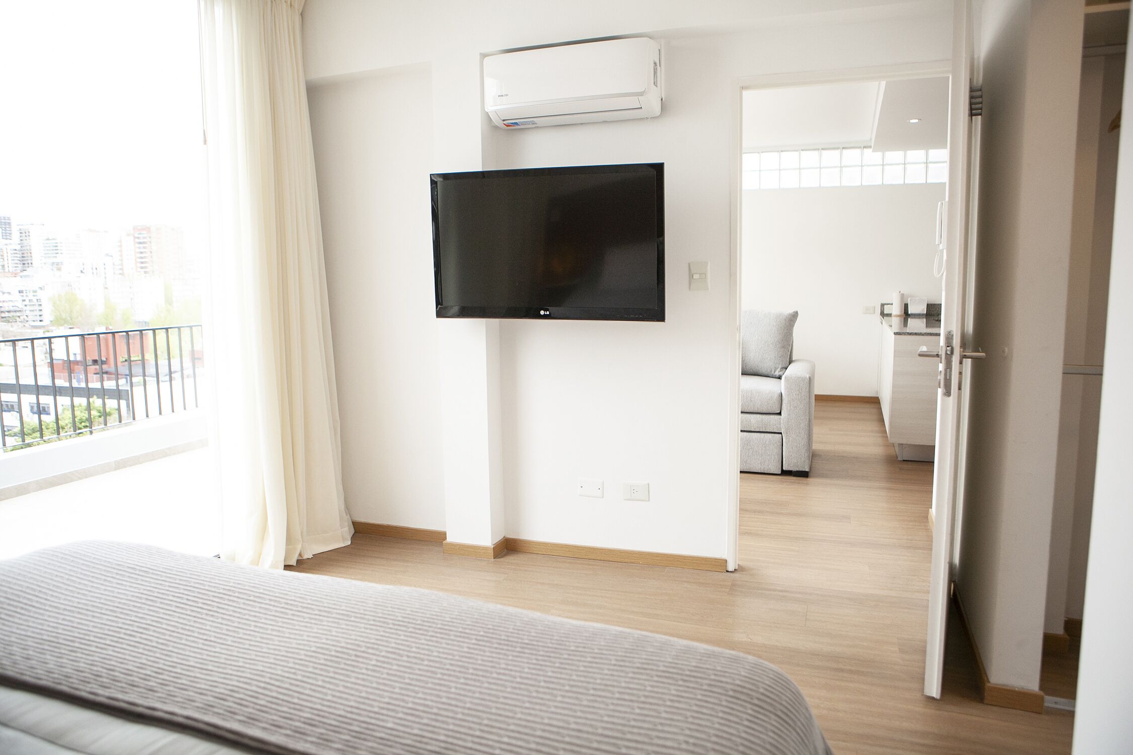 A modern hotel room with a flat-screen TV mounted on the wall, a bed with a striped cover, and a view of the city through the window.