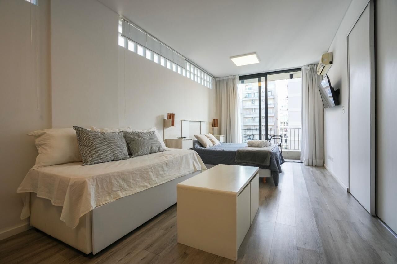 A modern hotel room with a large bed, a smaller bed, a bedside table, a TV mounted on the wall, and a view of the city through large windows.
