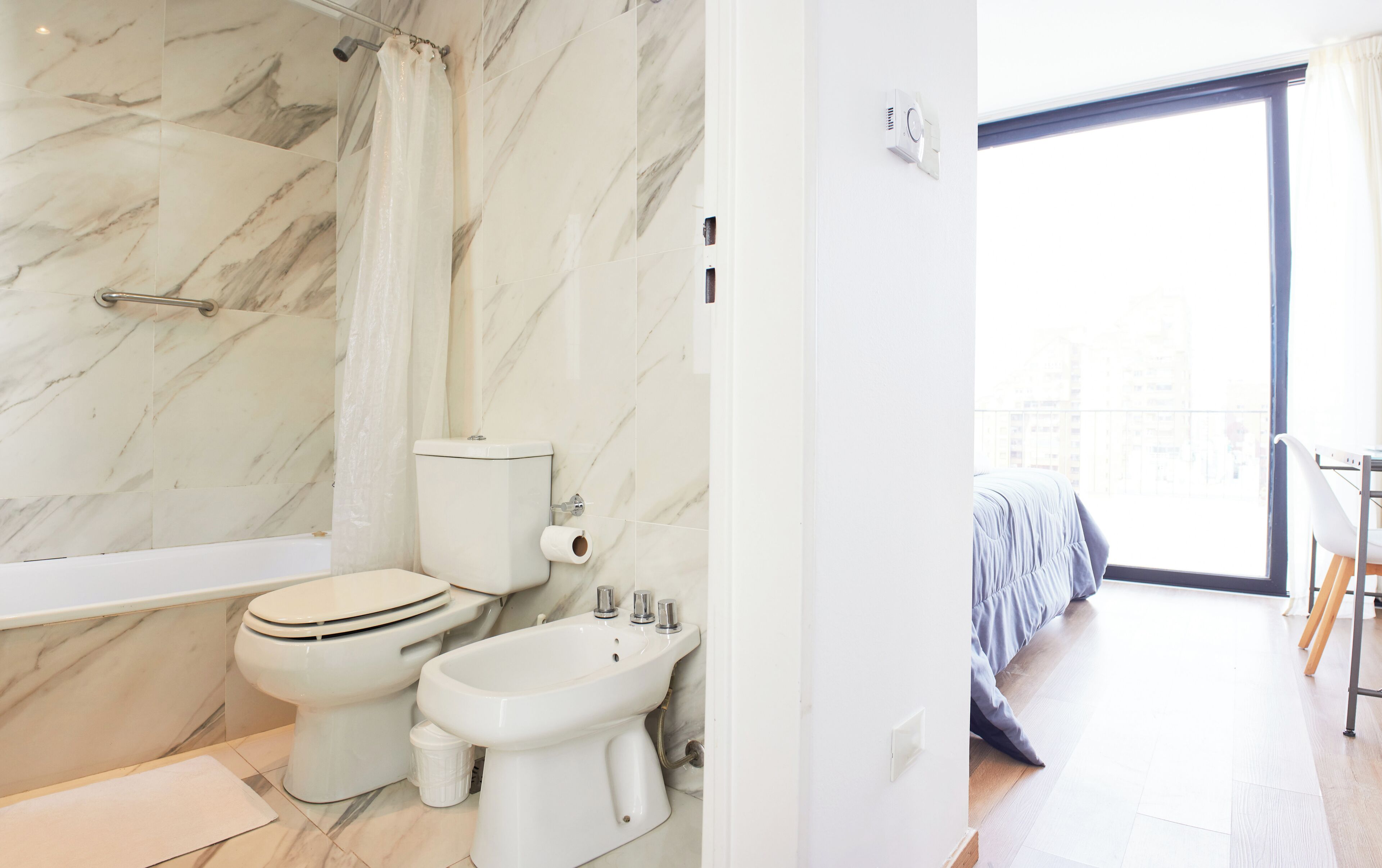 A modern bathroom with a bathtub, toilet, and bidet, marble walls, and a view of a balcony with a bed.