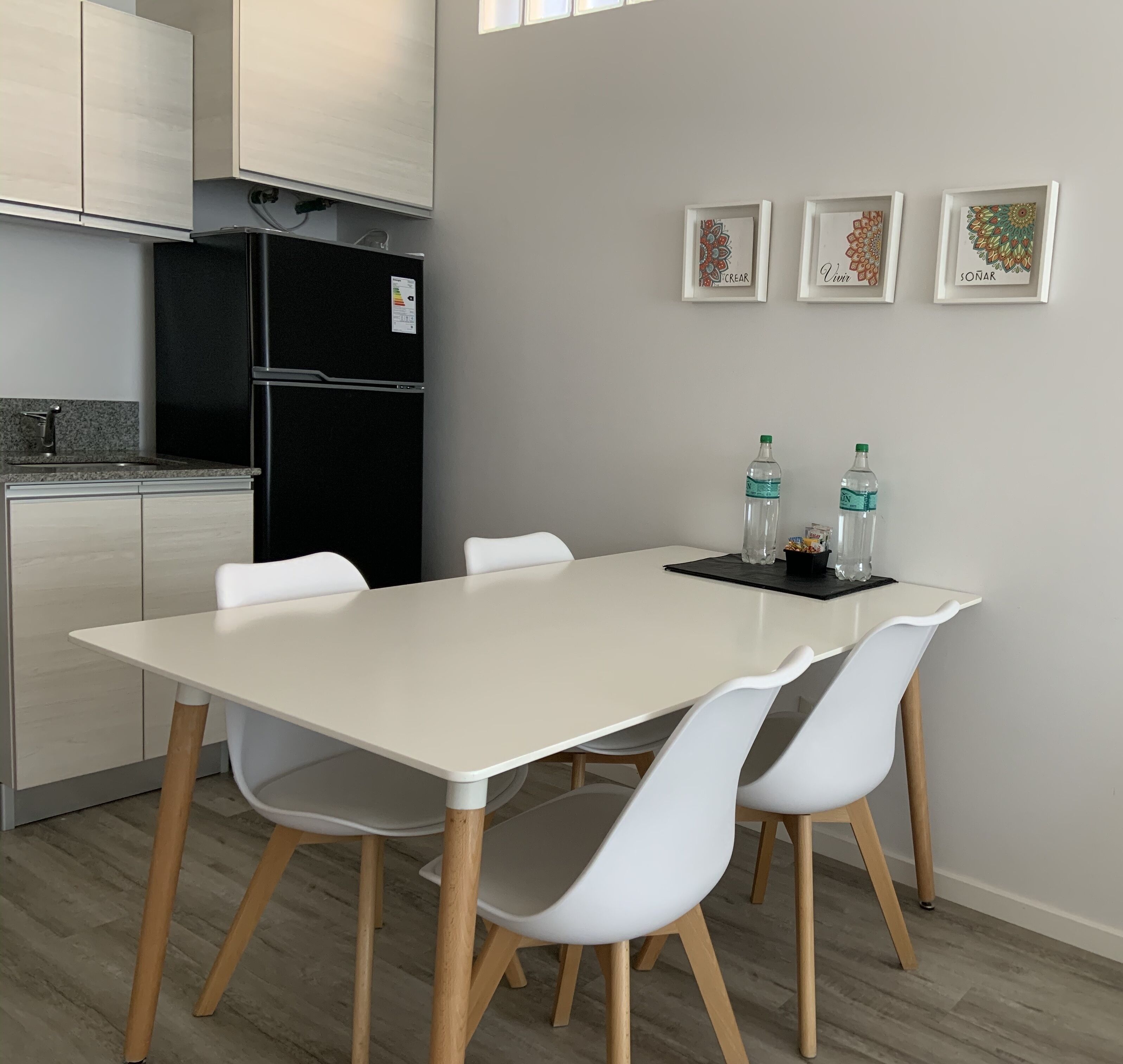 Une cuisine moderne avec une table et des chaises blanches, un réfrigérateur noir et des œuvres d’art encadrées accrochées au mur.