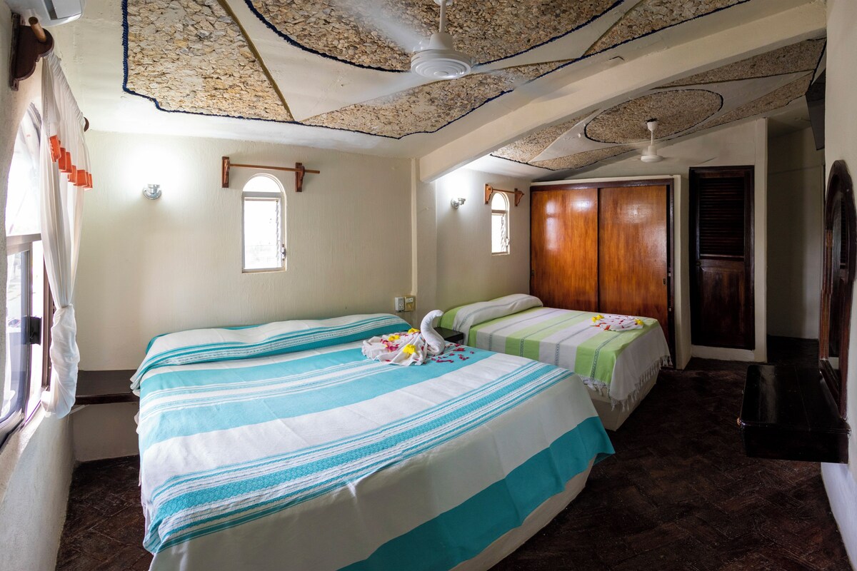 Quarto de hotel com duas camas, um guarda-roupa de madeira, um ventilador de teto e uma janela com cortinas.