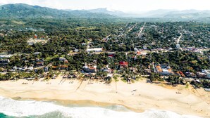 Vista aérea de un pueblo costero con playa, edificios residenciales y montañas al fondo.