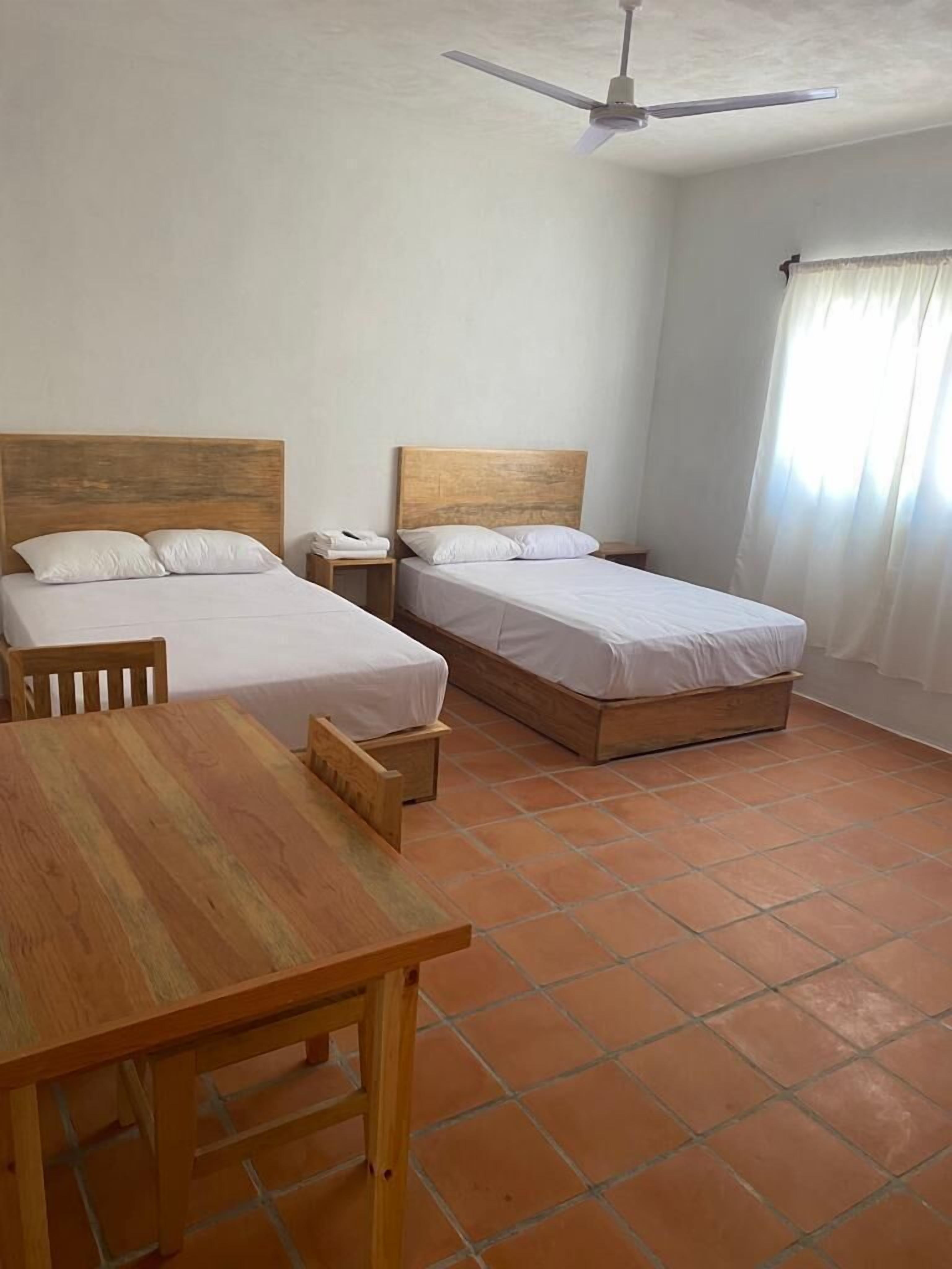 Een kamer met twee houten bedden, een houten tafel, een plafondventilator en een raam met gordijnen.