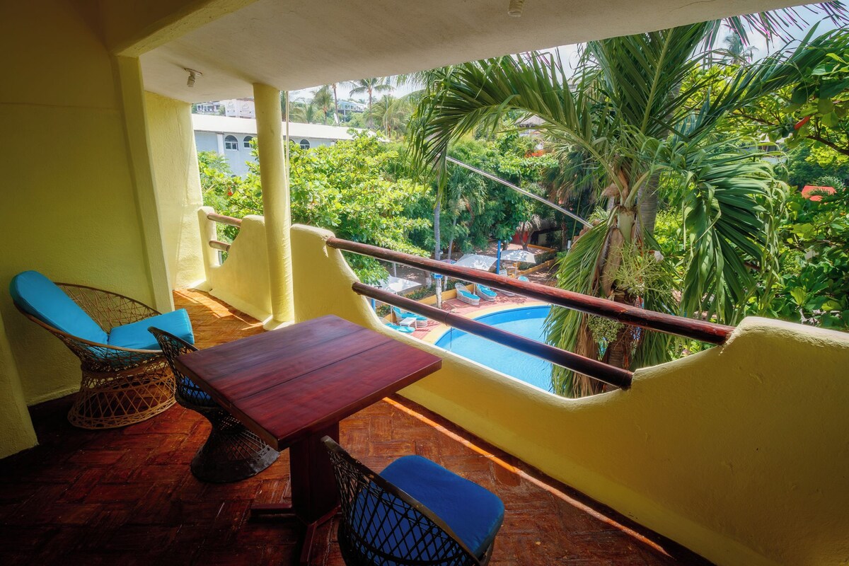 Uma varanda com mesa de madeira e cadeiras estofadas azuis, com vista para uma piscina e vegetação exuberante.