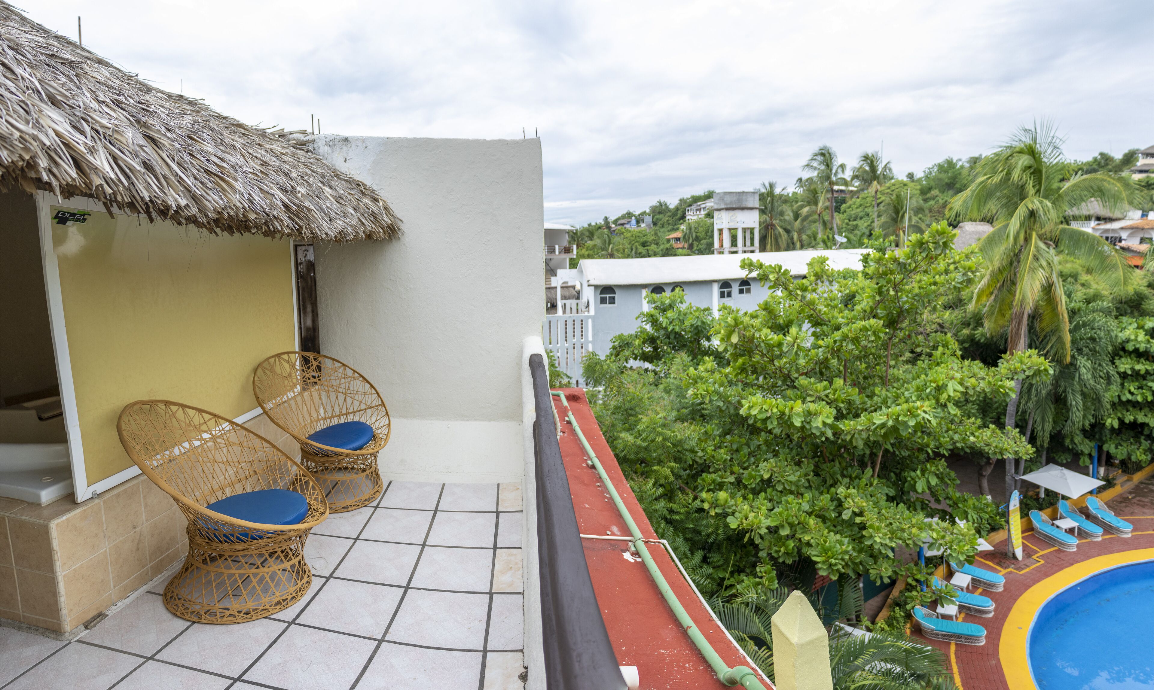 A balcony with wicker chairs, a thatched roof, and a view of a swimming pool and greenery.