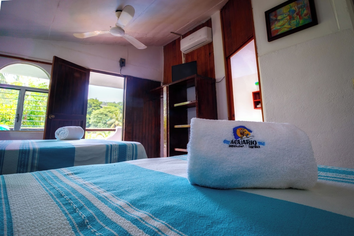 A room with two beds, a ceiling fan, an air conditioning unit, and a window with a view of greenery.