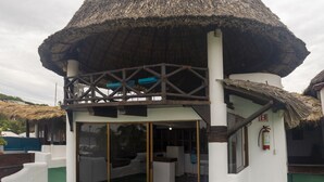 Una estructura con techo de paja y cerramiento de vidrio, paredes blancas y suelo verde.