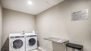 A laundry area with two front-loading washing machines, a metal shelf, and a trash bin. - Holiday Inn Express & Suites Tulsa East - Catoosa by IHG (Tulsa)