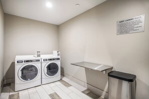 A laundry area with two front-loading washing machines, a metal shelf, and a trash bin. - Holiday Inn Express & Suites Tulsa East - Catoosa by IHG (Tulsa)