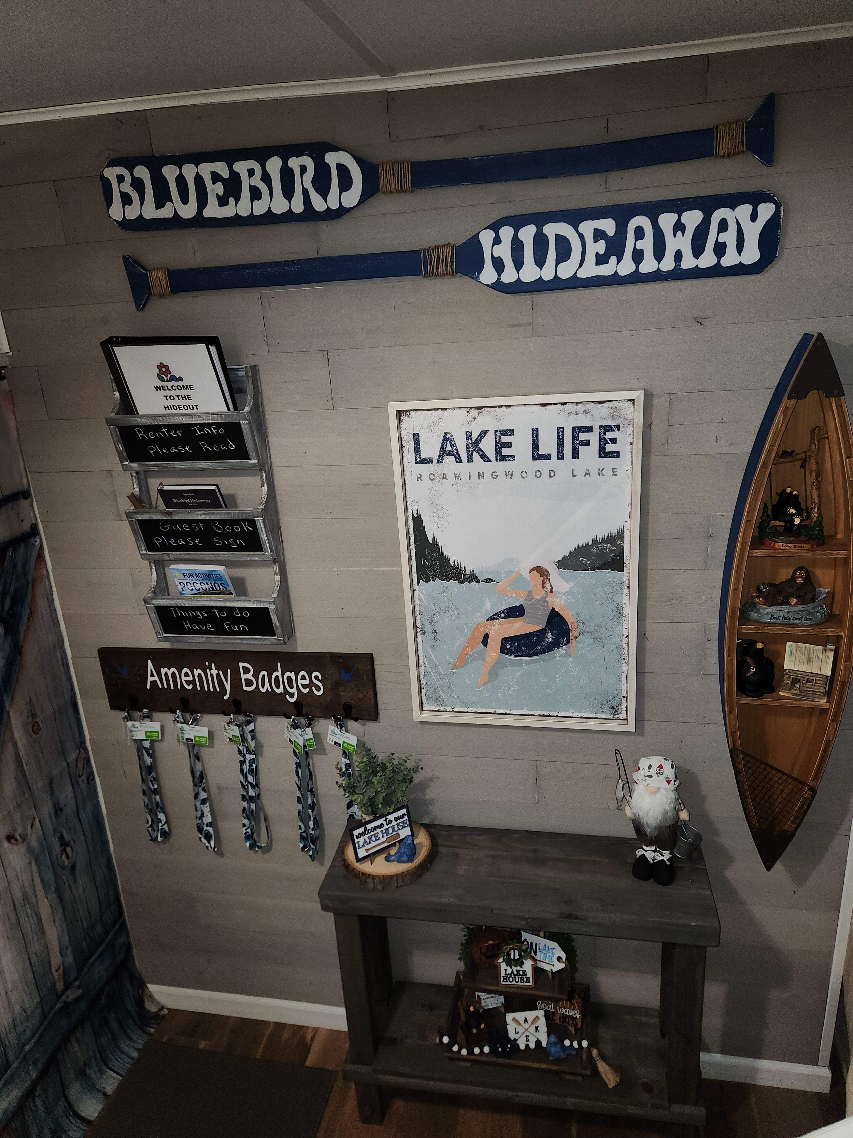 Een muur met verschillende borden en decoraties, waaronder een bord met 'Bluebird Hideaway', een poster met 'Lake Life' en een bord dat 'Gastenboek' aangeeft.
