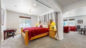 Um quarto espaçoso com uma cama grande, uma escrivaninha e uma cadeira, uma sala de estar separada com sofá e TV, e vista para árvores pela janela.