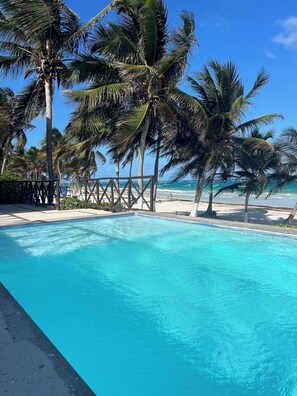 A clear blue pool surrounded by palm trees and a wooden deck, with a beach and ocean in the background. - El Paraiso Hotel Tulum (Tulum)