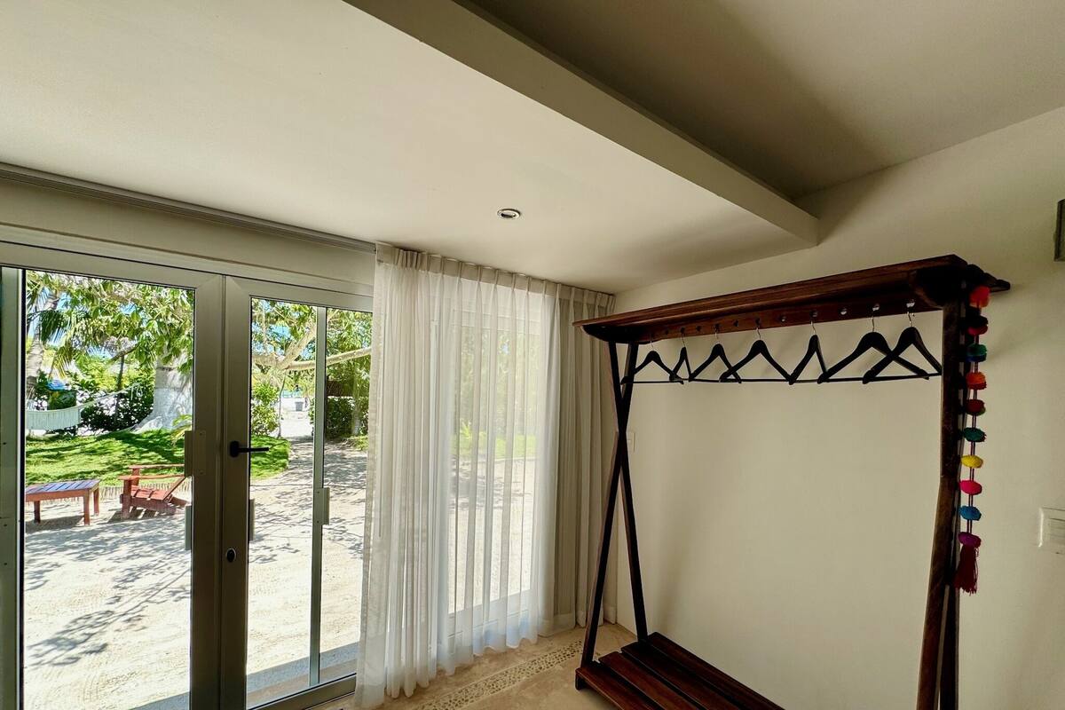 Ein Zimmer mit einer großen Glasschiebetür, die zu einem Außenbereich mit Hängematte und Stühlen führt. Es gibt ein Bett mit Holzrahmen und Baldachin sowie einen an der Wand befestigten Kleiderständer mit bunten Pompons.