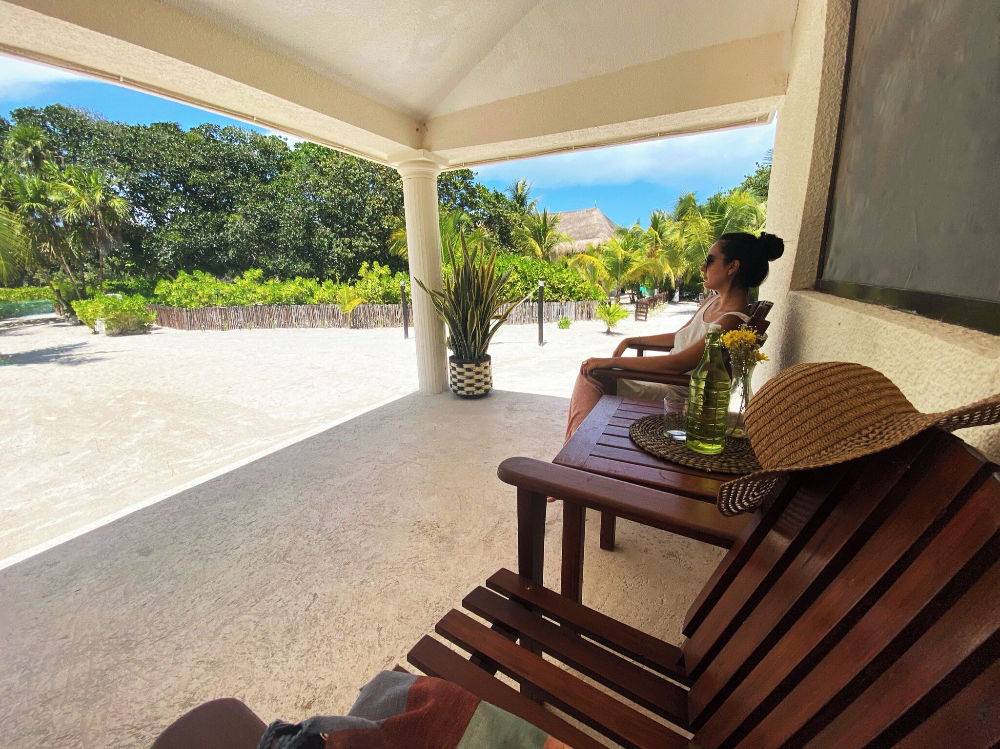 Un banco de madera con un cojín tejido y una botella de agua en un porche cubierto con vistas a una playa y palmeras.