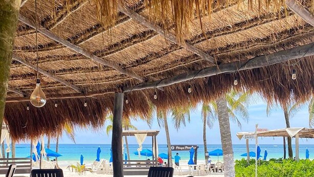 Zona pranzo in spiaggia con tavolo e sedie sotto un tetto di paglia, con vista sull'oceano.