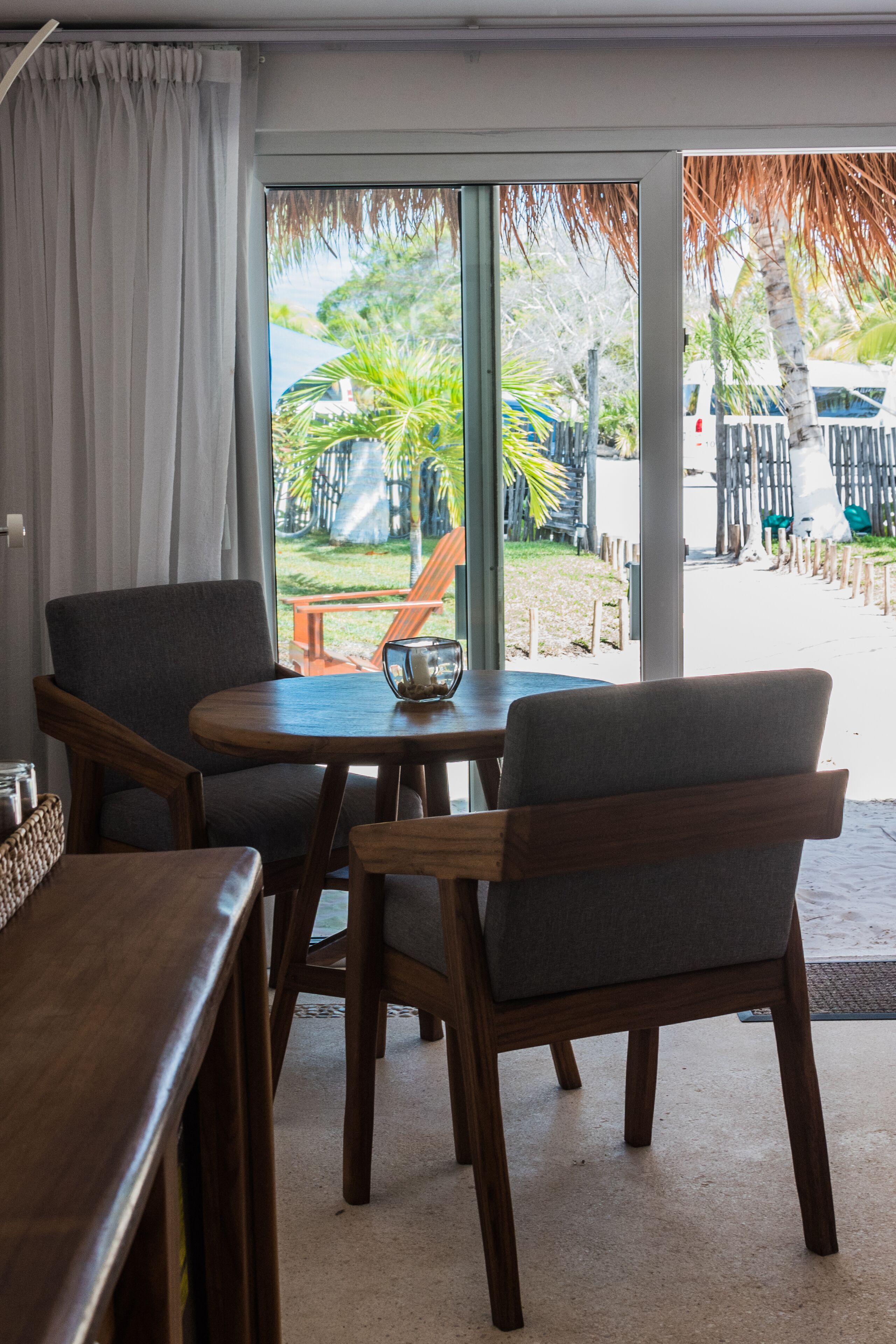 Habitación con una mesa y sillas de madera, vista a un jardín con palmeras y una playa visible a través de la ventana.