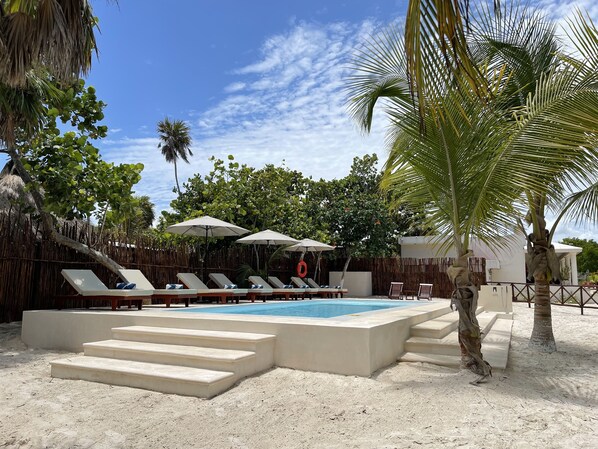 A pool area with lounge chairs and umbrellas, surrounded by palm trees and a sandy beach.