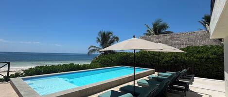 A pool area with lounge chairs and an umbrella, overlooking a beach and ocean.