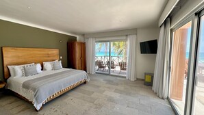 Una camera da letto con un letto grande, una testata in legno, una TV fissata al muro e vista sulla spiaggia tramite una porta scorrevole in vetro.