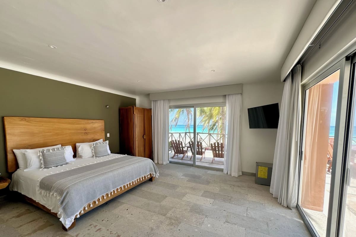 Ein Schlafzimmer mit einem großen Bett, einem hölzernen Kopfteil, einem an der Wand montierten Fernseher und Blick auf den Strand durch eine Schiebetür.