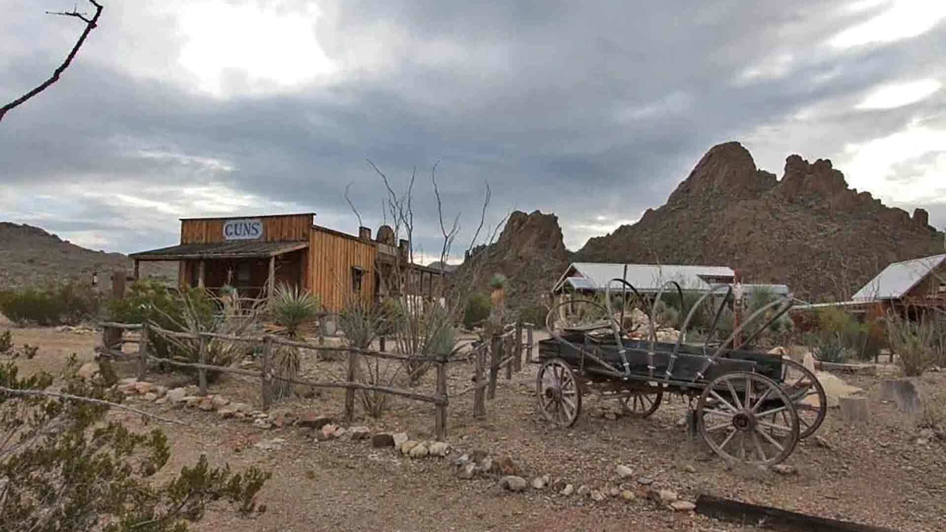 A rustic building labeled 'GUNS' with a wooden exterior, surrounded by sparse vegetation and a rugged mountain backdrop.