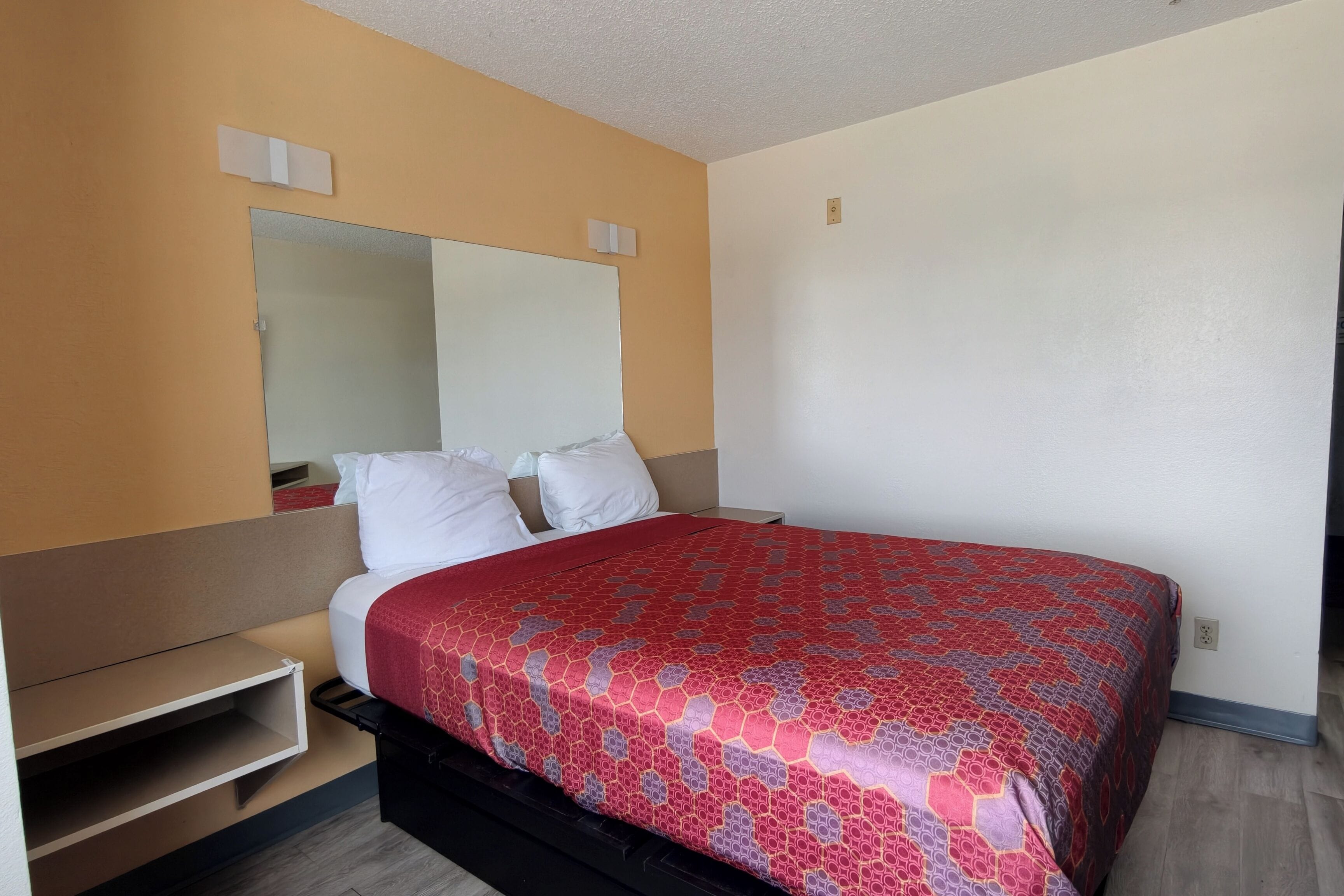 Hotellihuone, jossa on sänky, yöpöytä, peili ja seinään integroitu hylly.
