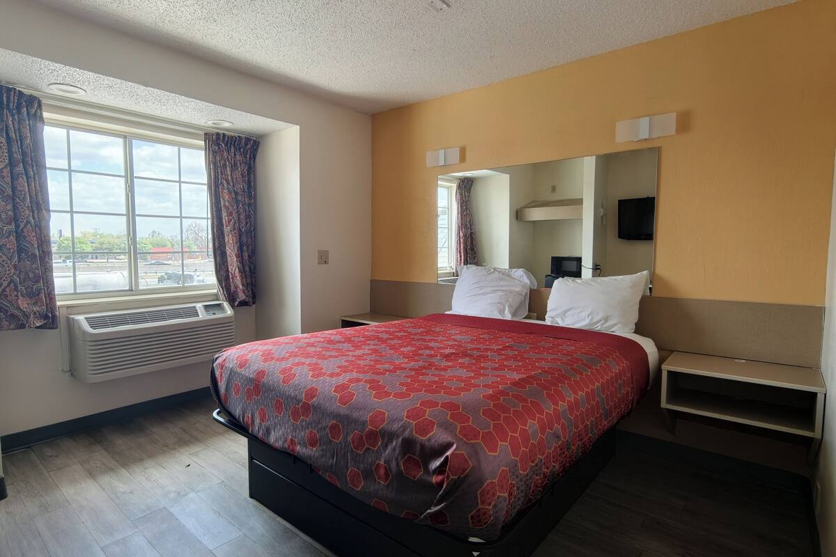 Ein Hotelzimmer mit Bett, einem Vorhangfenster, einem Heizkörper, einem Spiegel und einem Fernseher.