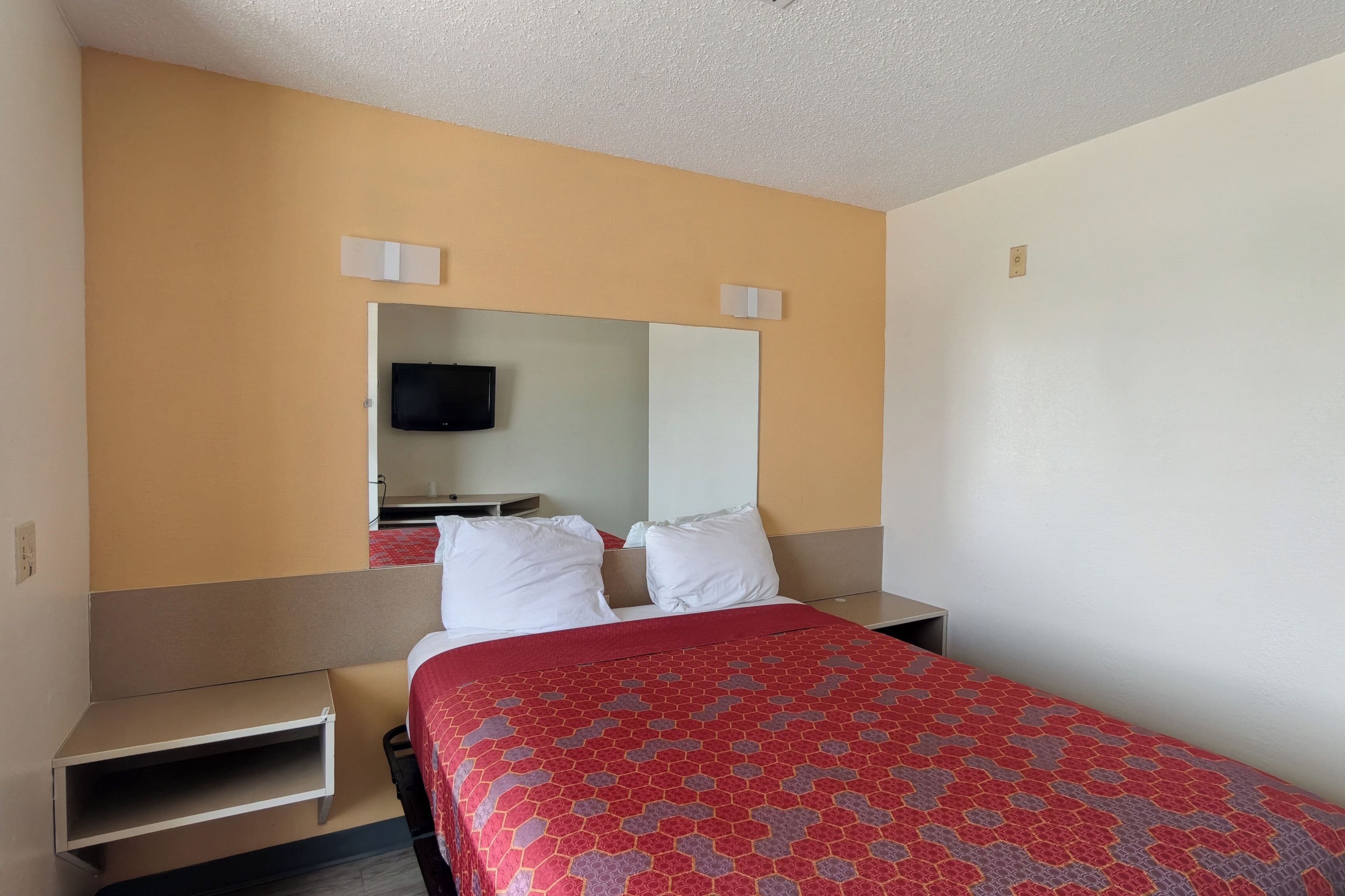 A hotel room with a bed, a TV, a mirror, and a small shelf.