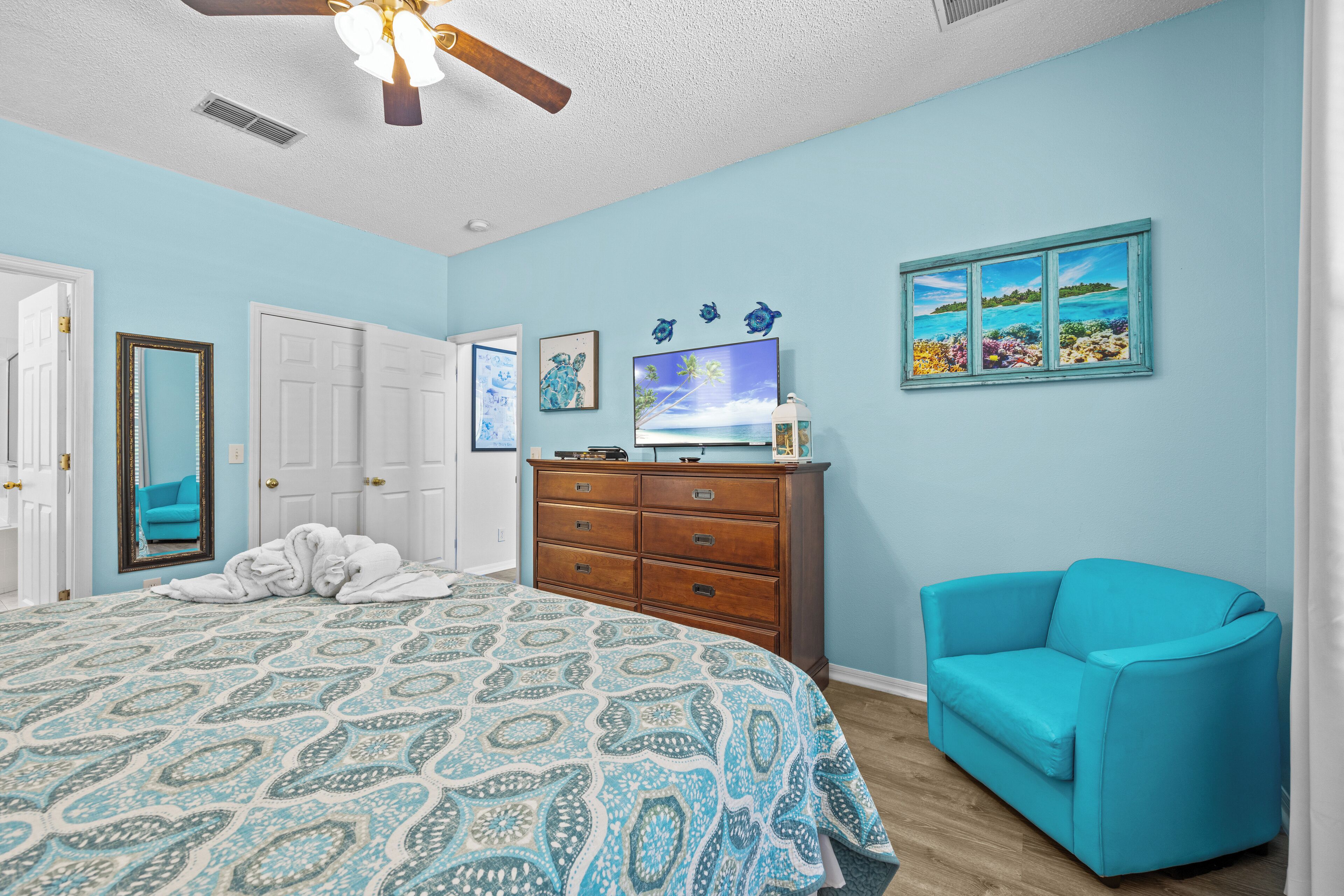 Un dormitorio con cama, cómoda, sillón azul turquesa y televisor sobre la cómoda.