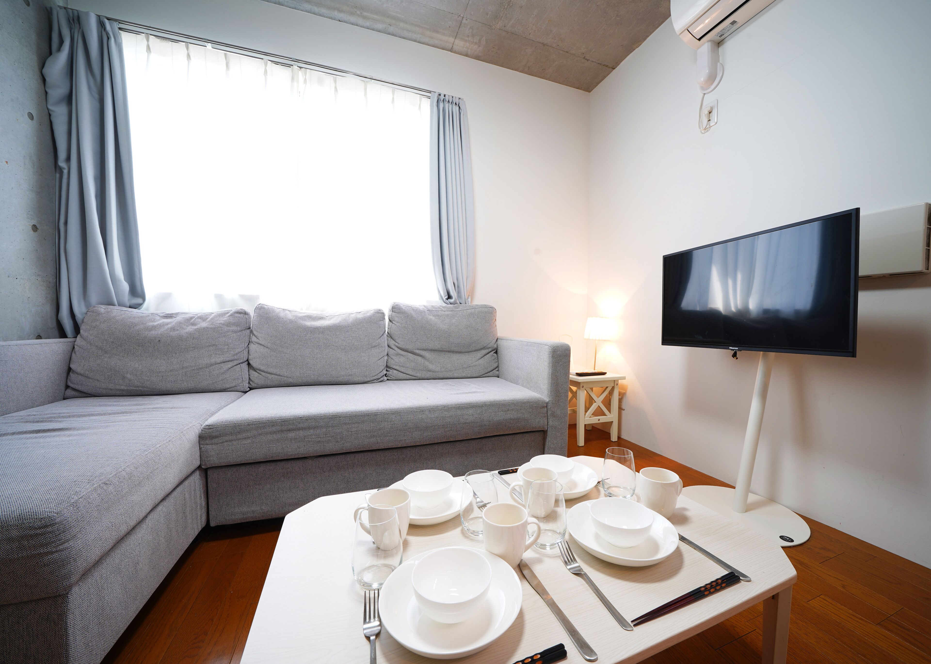 A modern living room with a grey sofa, a dining table set with white plates, glasses, and cutlery, a flat-screen TV mounted on the wall, and a lamp on a side table.