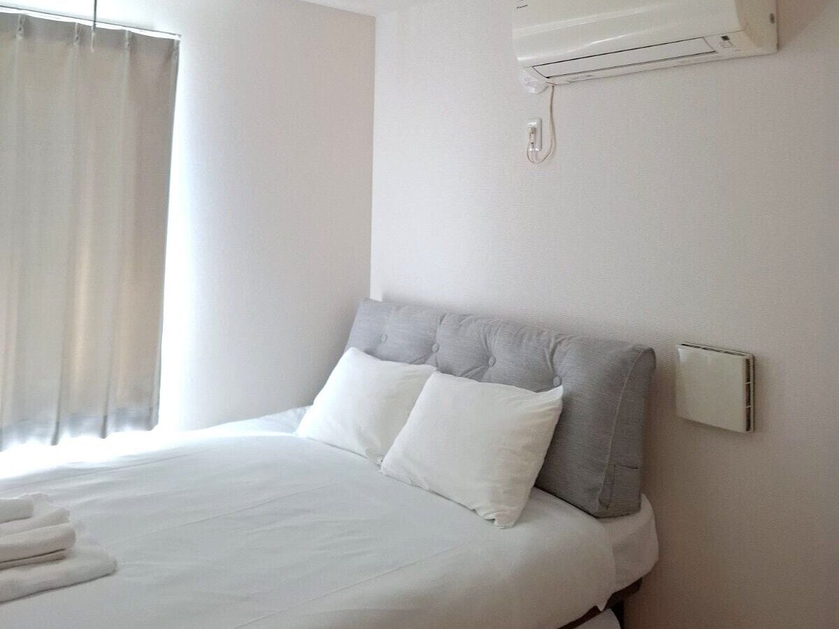 Habitación de hotel con una cama, ropa de cama blanca, cabecera gris y un equipo de aire acondicionado en la pared.