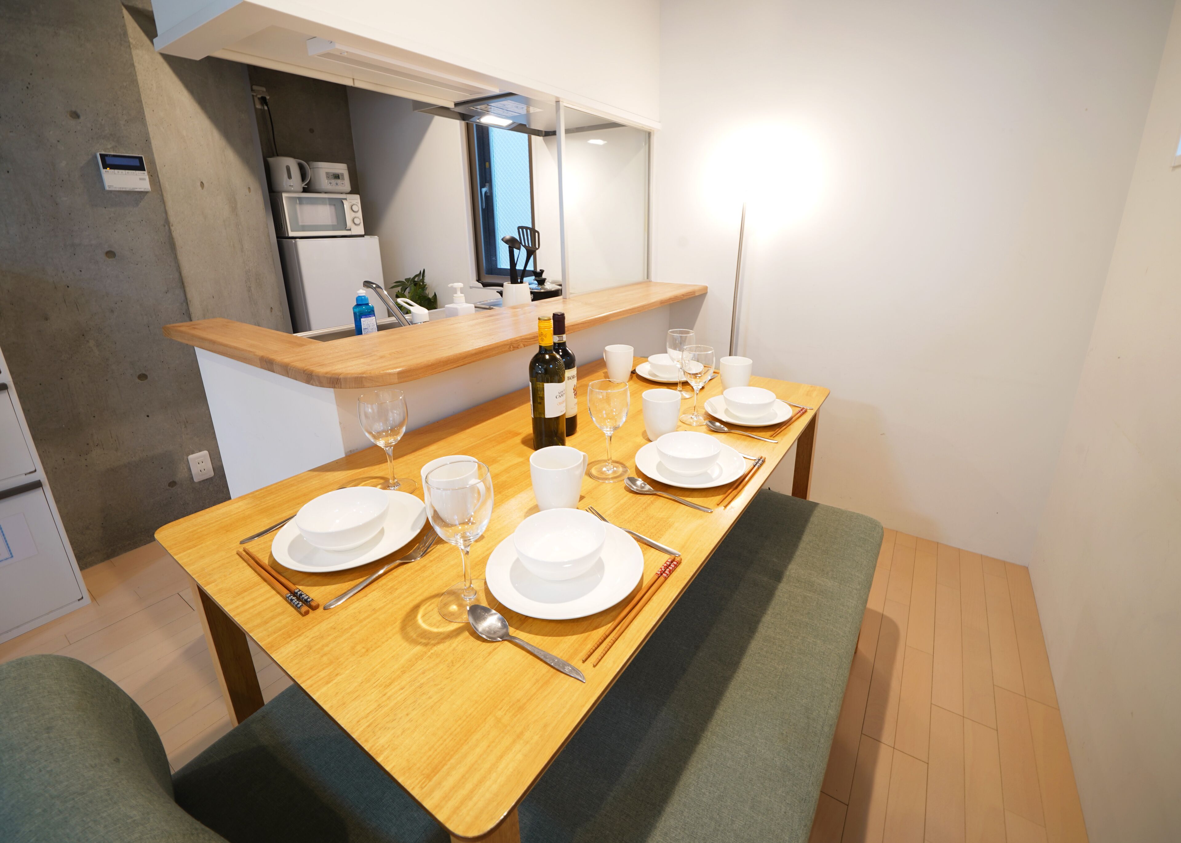 Comedor con mesa de madera dispuesta para cuatro, incluyendo platos, vasos y cubiertos. Se ve una botella de vino y un microondas al fondo.
