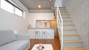 A modern living room with a grey sofa, a wooden staircase, and a kitchen area. - Laffitte Hirai Condominium Hotel (Tokyo)