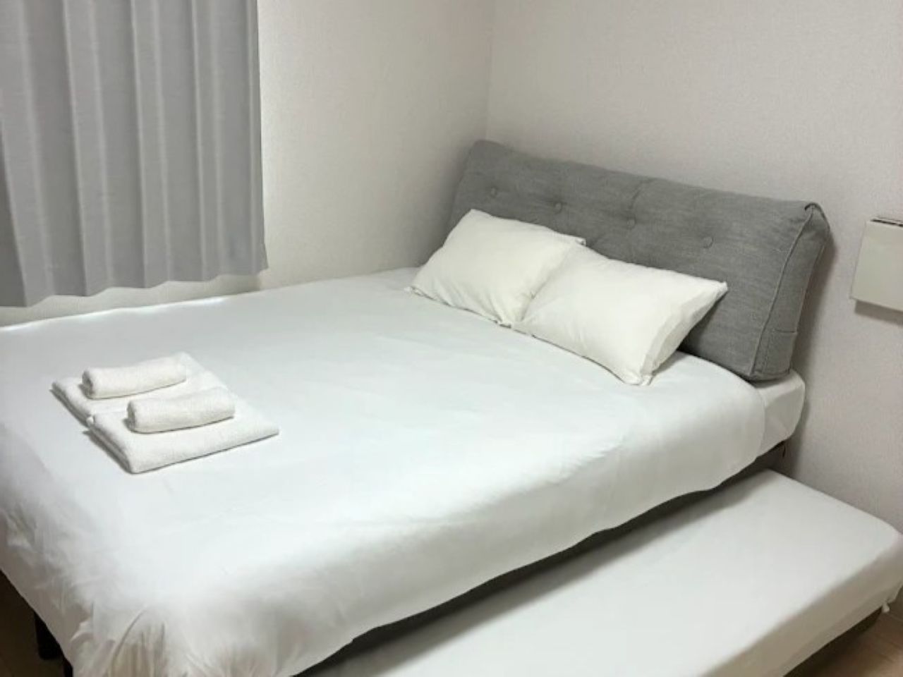 A single bed with a grey headboard, white bedding, and two rolled towels.
