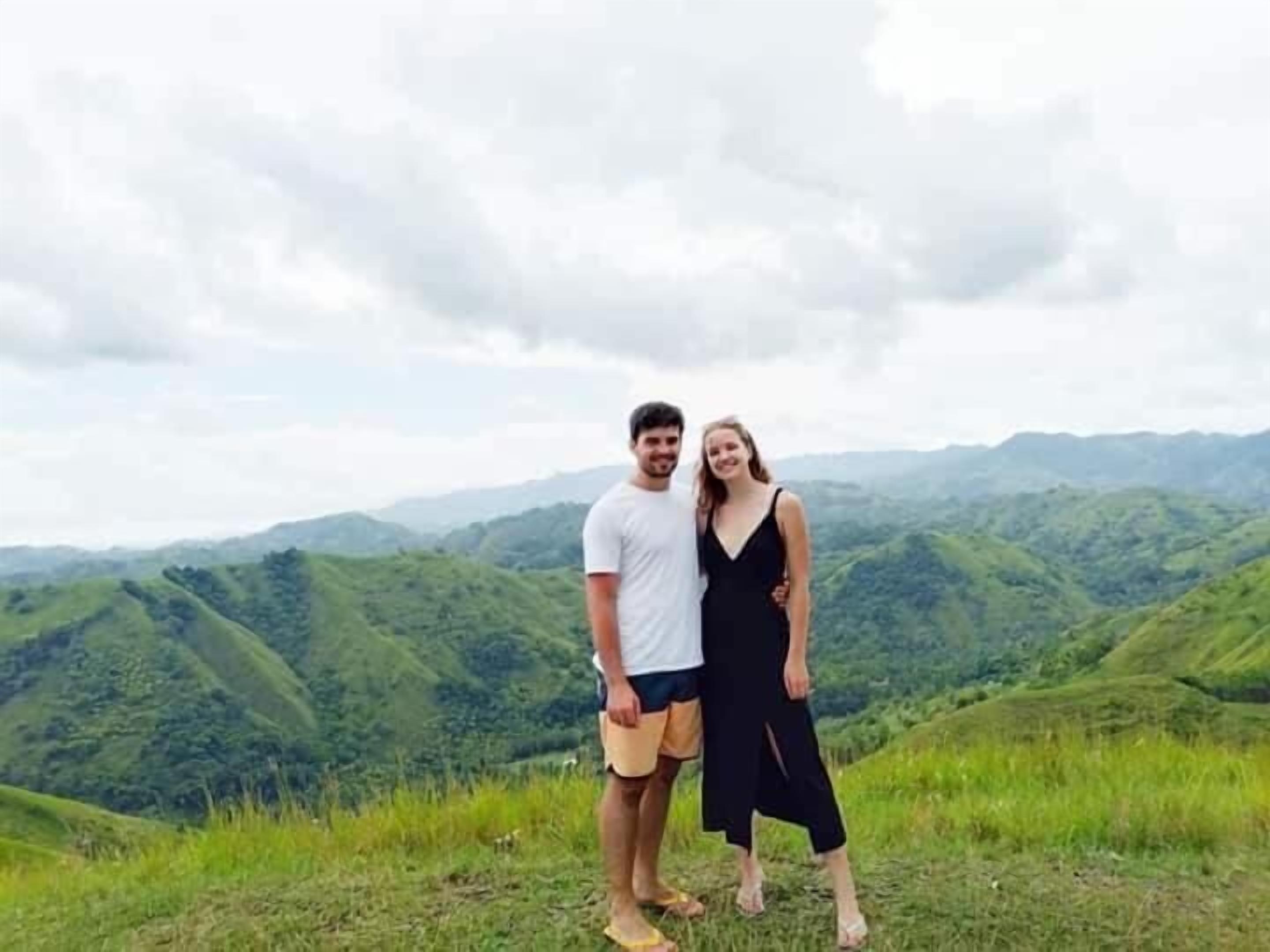 A couple standing on a grassy hill with a mountainous landscape in the background.
